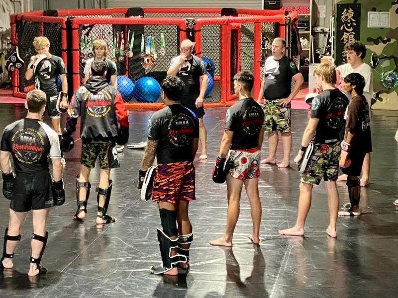 A kickboxing class gathered with pads and shin guards beside the cage