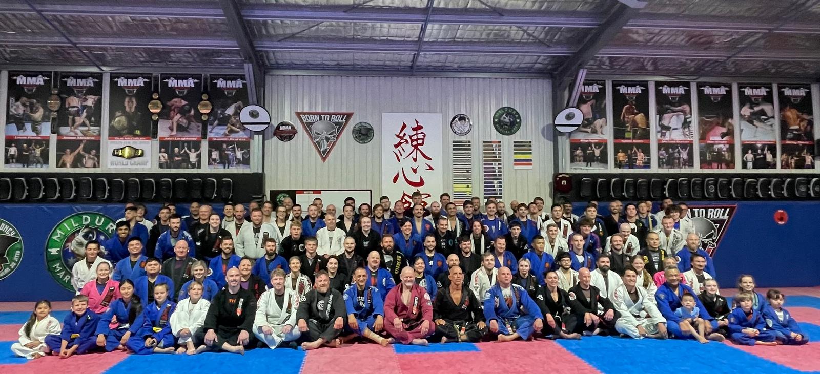 Kids and adults in their gis sitting and standing together on the mats under the Born To Roll banner and the Renshinkan sign