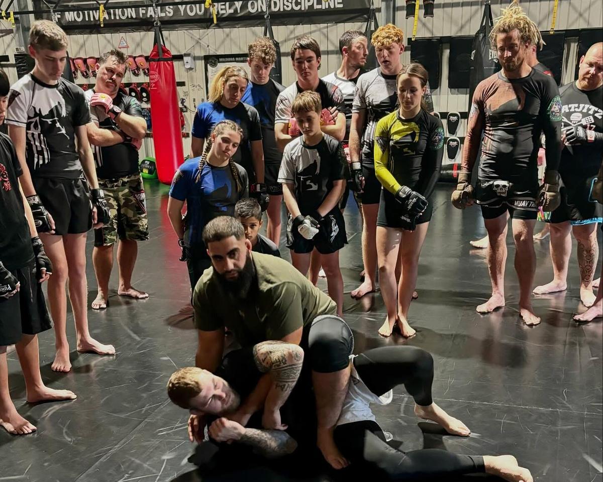 A coach demonstrating a pin during an MMA class while kids and adults watch closely